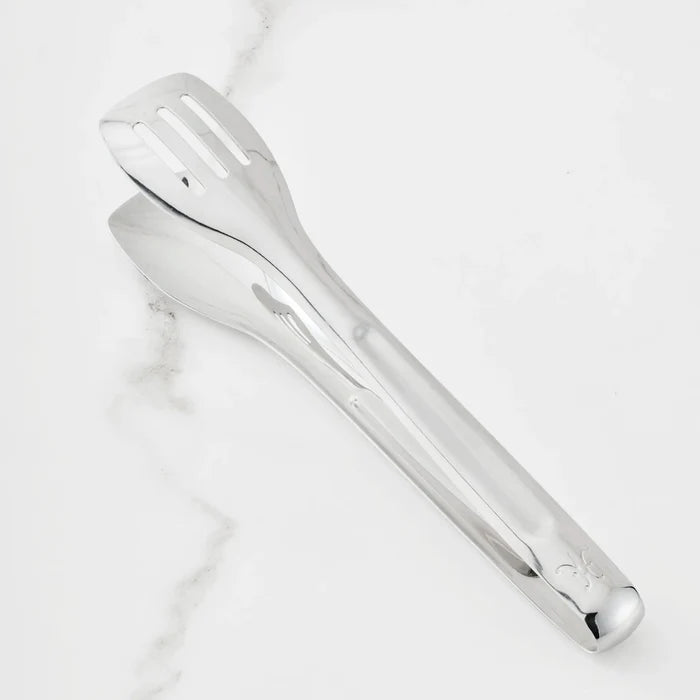 A pair of 10-inch Stainless Steel Serving Tongs from the Hestan Provisions® collection rests on a white marble surface. One side features a slotted, flat end while the other has a spoon-shaped end, and the handle is finished with a small embossed design near the tip—an elegant addition to any chef’s kitchen tools.