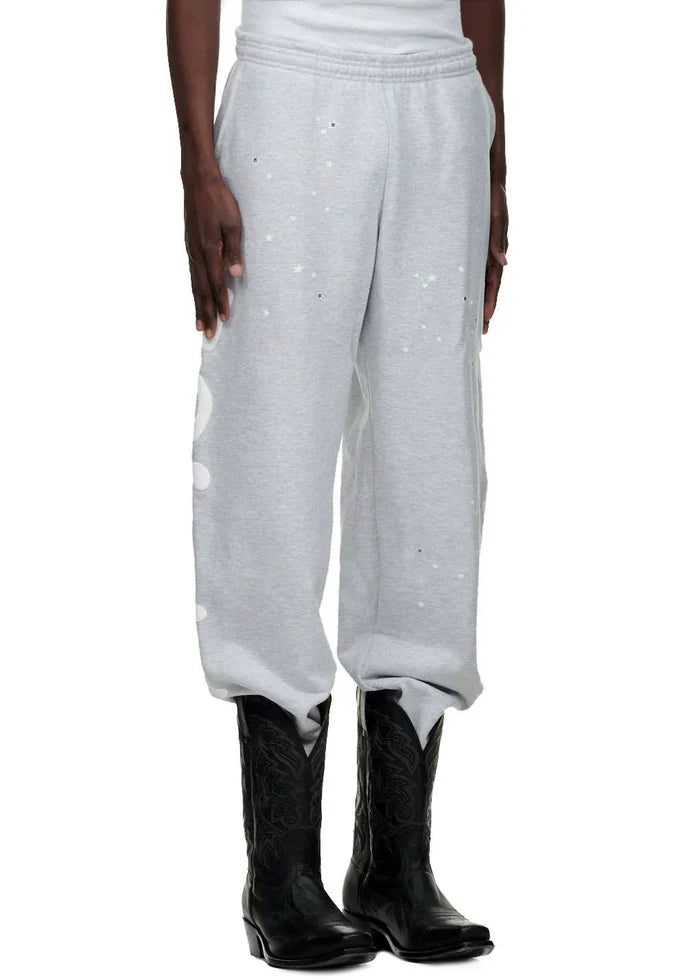 A person wearing 100% Cotton Sp5der Sweatpants for Men – Grey Streetwear Jogger Pants with Graphic Print, paired with a white top and black cowboy boots, showcases a casual streetwear look against a white background.