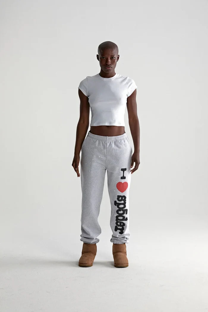 A person faces forward in a fitted white crop top, brown boots, and light grey Sp5der jogger pants featuring “I Love Sp5der” graphic, relaxed fit—showcasing casual streetwear style against a plain white backdrop.