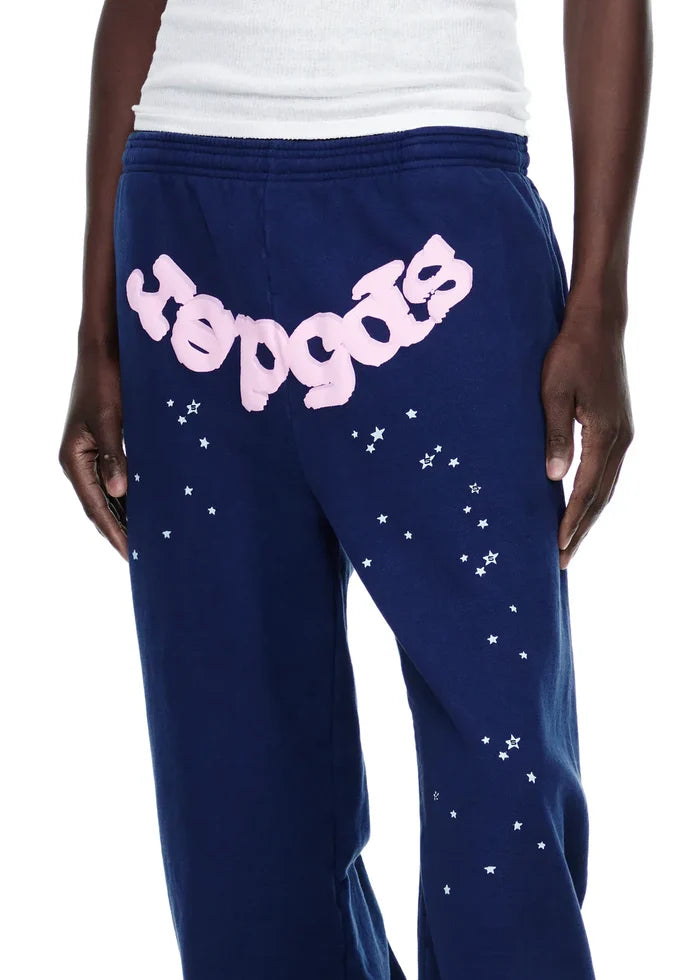 A person wearing 100% Cotton Sp5der Sweatpants—unisex navy blue joggers with scattered white star graphics and a pink logo—stands with one hand at their side.