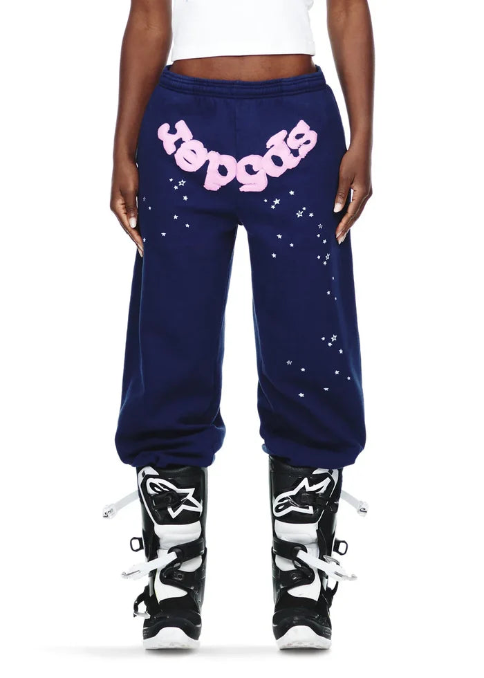 A person wears 100% Cotton Sp5der Sweatpants—unisex navy blue jogger pants with a pink logo and star graphics, relaxed fit—paired with black and white motocross boots. Their torso is cropped out of the image.