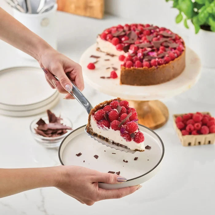 Using the Hestan Provisions® 10.5-inch Stainless Steel Pie & Cake Server, a person places a slice of raspberry-topped cheesecake with chocolate shavings onto a plate. The rest of the cheesecake remains on a round stand nearby, while a bowl of raspberries and other Hestan Provisions® Serving Tools sit in the background on a white countertop.