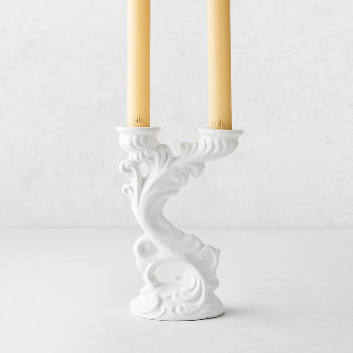 Assisi white ceramic candlestick with ornate scrollwork, holding two yellow taper candles, 7.5 inches tall.