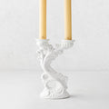 Assisi white ceramic candlestick with ornate scrollwork, holding two yellow taper candles, 7.5 inches tall.