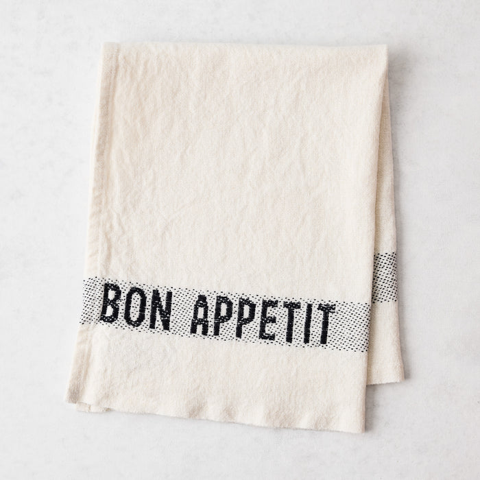 Off-white linen napkin with "BON APPETIT" in black lettering, folded on a white surface.