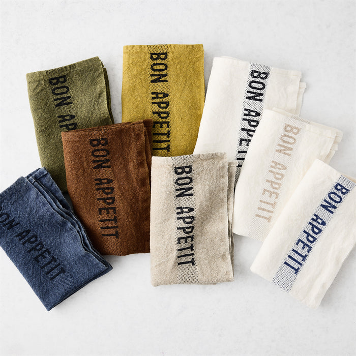 Folded linen napkins in various colors, each with "BON APPETIT" text.