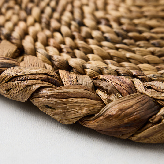 Close-up of a round, honey-brown water hyacinth placemat featuring a detailed braided border and woven texture.
