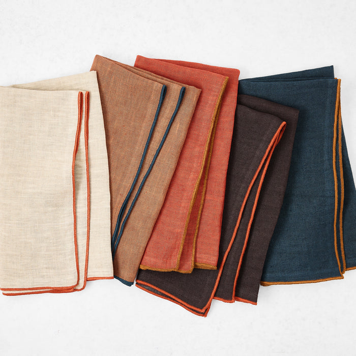 Set of square linen napkins in assorted earth tones with contrasting embroidered tipped edges.
