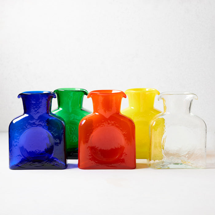 Five square glass pitchers in blue, green, red, yellow, and clear, with dual spouts and side dips.