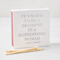 Square white matchbox with holographic text reading It's Hard to be a Diamond in a Rhinestone World by Dolly Parton.