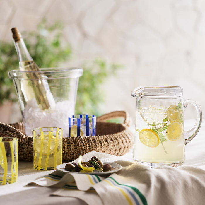 Clear Oxford Glass Ice Bucket with champagne bottle, pitcher of lemonade, and striped glasses on a woven tray.