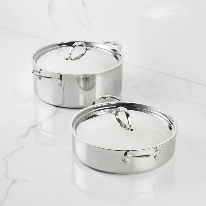Two Polished Clad Stainless Steel Rondeaus with lids from the ProBond Luxe® collection are placed on a white marble countertop. Each rondeau features metal handles and a polished, reflective surface, while the matching lids are topped with elegant looped handles.