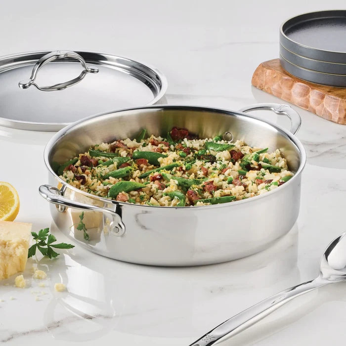 A ProBond Luxe® Polished Clad Stainless Steel Rondeau with Lid sits on a marble countertop, filled with cooked rice, green beans, and bits of sausage. Nearby are a large spoon, plates, a lemon wedge, cheese, parsley, and the lid.