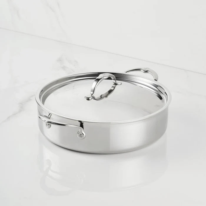A ProBond Luxe® Polished Clad Stainless Steel Rondeau with Lid rests on a white marble surface. The lid features a loop handle, and the rondeau is designed with two side handles. Both the surface and the cookware shimmer beautifully in the light.
