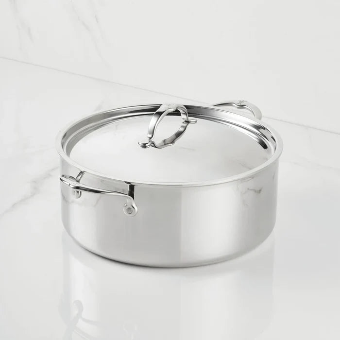 A ProBond Luxe® Polished Clad Stainless Steel Rondeau with Lid rests on a white marble countertop. The rondeau showcases sturdy handles on both the sides and lid, while its polished surface mirrors the pristine surroundings.