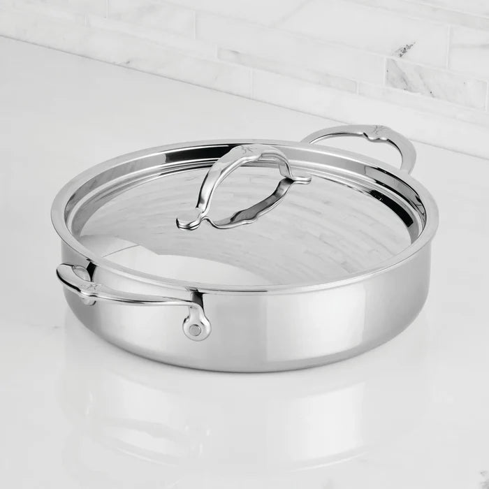 A ProBond Luxe® Polished Clad Stainless Steel Rondeau with Lid sits on a white marble countertop, set against a backdrop of tile. Part of ProBond Luxe®'s clad stainless steel cookware collection, the rondeau features two handles and boasts a shiny, reflective finish.
