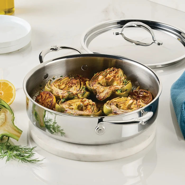 A ProBond Luxe® Polished Clad Stainless Steel Rondeau with Lid, filled with cooked, halved artichokes, sits on a marble countertop. The nearby lid, along with a lemon slice, herbs, and a blue cloth, highlights the elegance of clad stainless steel cookware in the modern kitchen.