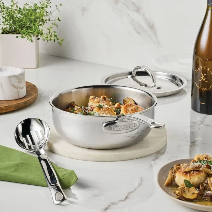 A polished stainless steel pan from the ProBond Luxe® Polished Clad Stainless Steel Signature Set, 7-Piece sits on a marble surface filled with baked chicken and vegetables. Nearby are a slotted spoon, green napkin, plated meal, wine bottle, and salt container. In the background, the tri-ply clad cookware lid—crafted from induction-suitable stainless steel—showcases the premium quality of ProBond Luxe®.