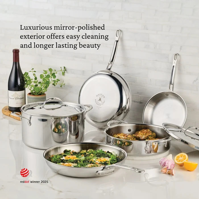 A gleaming ProBond Luxe® Polished Clad Stainless Steel Saucier Pan, 2-Quart, sits on a white countertop, filled with delicious cooked food. Surrounding the pan are fresh herbs, lemons, and a wine bottle. A nearby text highlights the saucier’s polished exterior and easy-to-clean benefits.